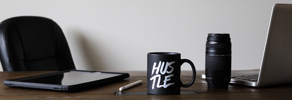 Image of a work desk featuring a tablet, laptop, camera lens, pen and coffee mug with the word "HUSTLE" printed on it.