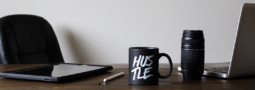 Image of a work desk featuring a tablet, laptop, camera lens, pen and coffee mug with the word "HUSTLE" printed on it.