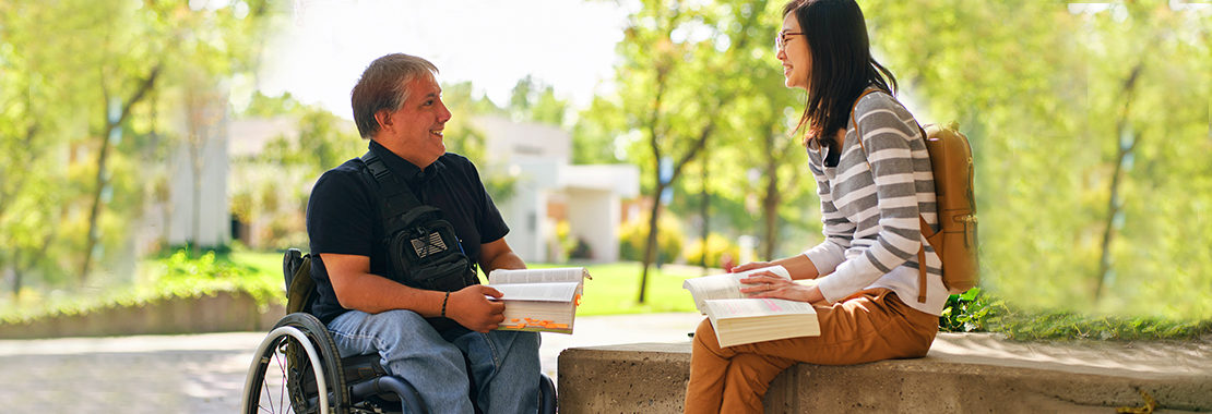 Two students talking with one another outside, one sitting in a wheel chair and one on a bench