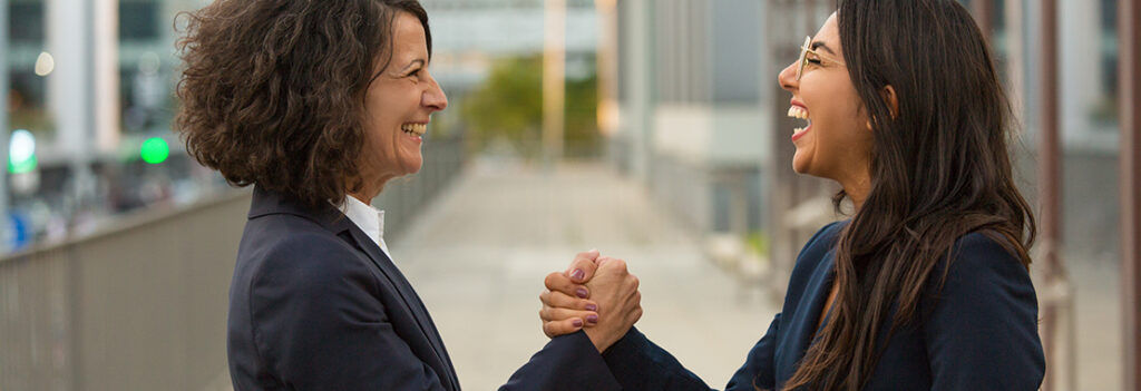 Photo of two women in business attire grasping hands and smiling.