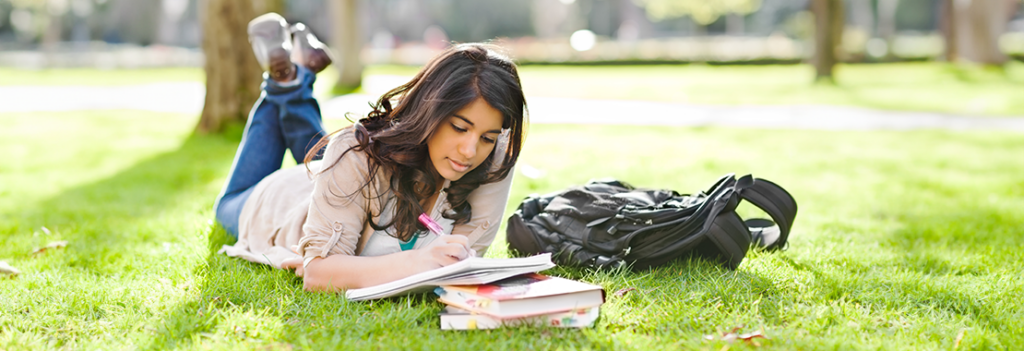 Photo of a female student laying in the grass studying a textbook.