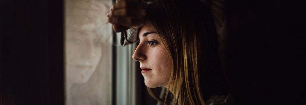 A young woman looks out a window, while her reflection shows