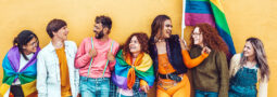 A collection of young people standing in front of a pride flag, wearing rainbow flags and colors.