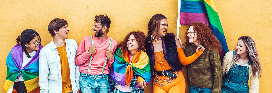 A collection of young people standing in front of a pride flag, wearing rainbow flags and colors.