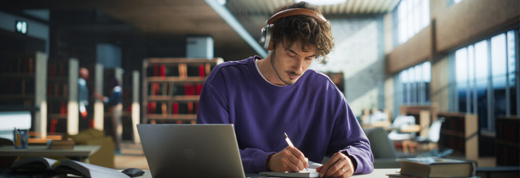 A male student with headphones in a library, sits while writing
