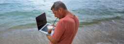 Man stands and works on laptop at the beach while sipping iced coffee