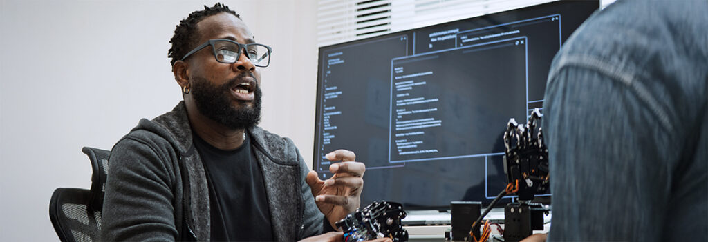 An individual sits by their desktop computer with robotic hands set nearby.