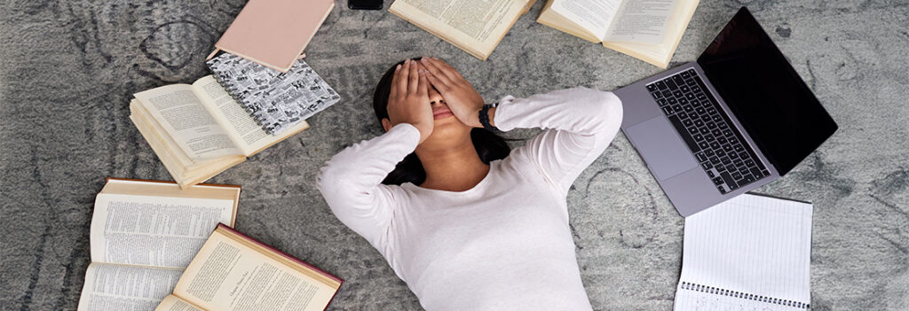 An student struggling with mental wellbeing, lies on the ground surrounded by open books, her hands covering her face.