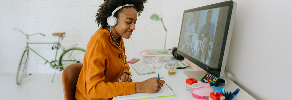 A woman wearing headphones write in her notepad and works at her desktop computer.