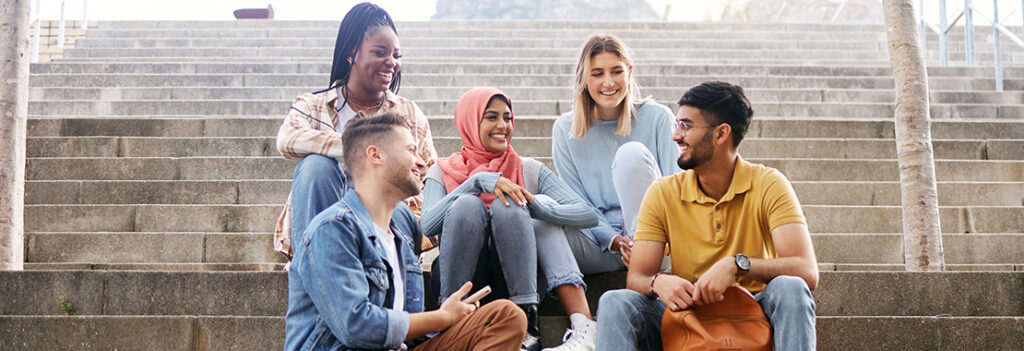 Five college peers smile and connect with each other while sitting on concrete steps.