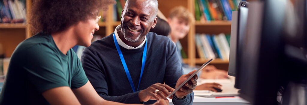 Two people sitting at a desk in a library or study area, looking at a tablet together. Bookshelves are visible in the background, and a computer monitor is in the foreground.