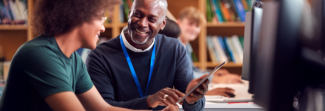 Two people sitting at a desk in a library or study area, looking at a tablet together. Bookshelves are visible in the background, and a computer monitor is in the foreground.
